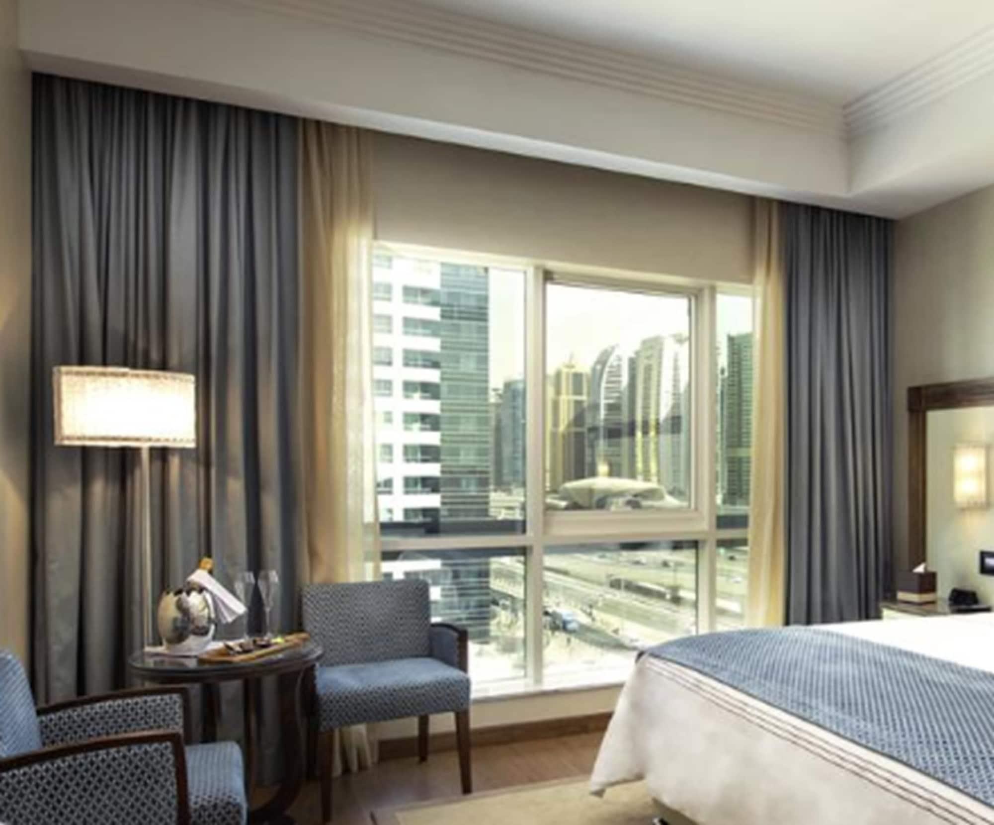 Stella Di Mare Dubai Marina Hotel Esterno foto The image shows a hotel room with a modern and stylish design. There are large windows that allow natural light to flood the space, revealing a view of city buildings outside. The room features a bed with crisp linens, a bedside table with a lamp, an
