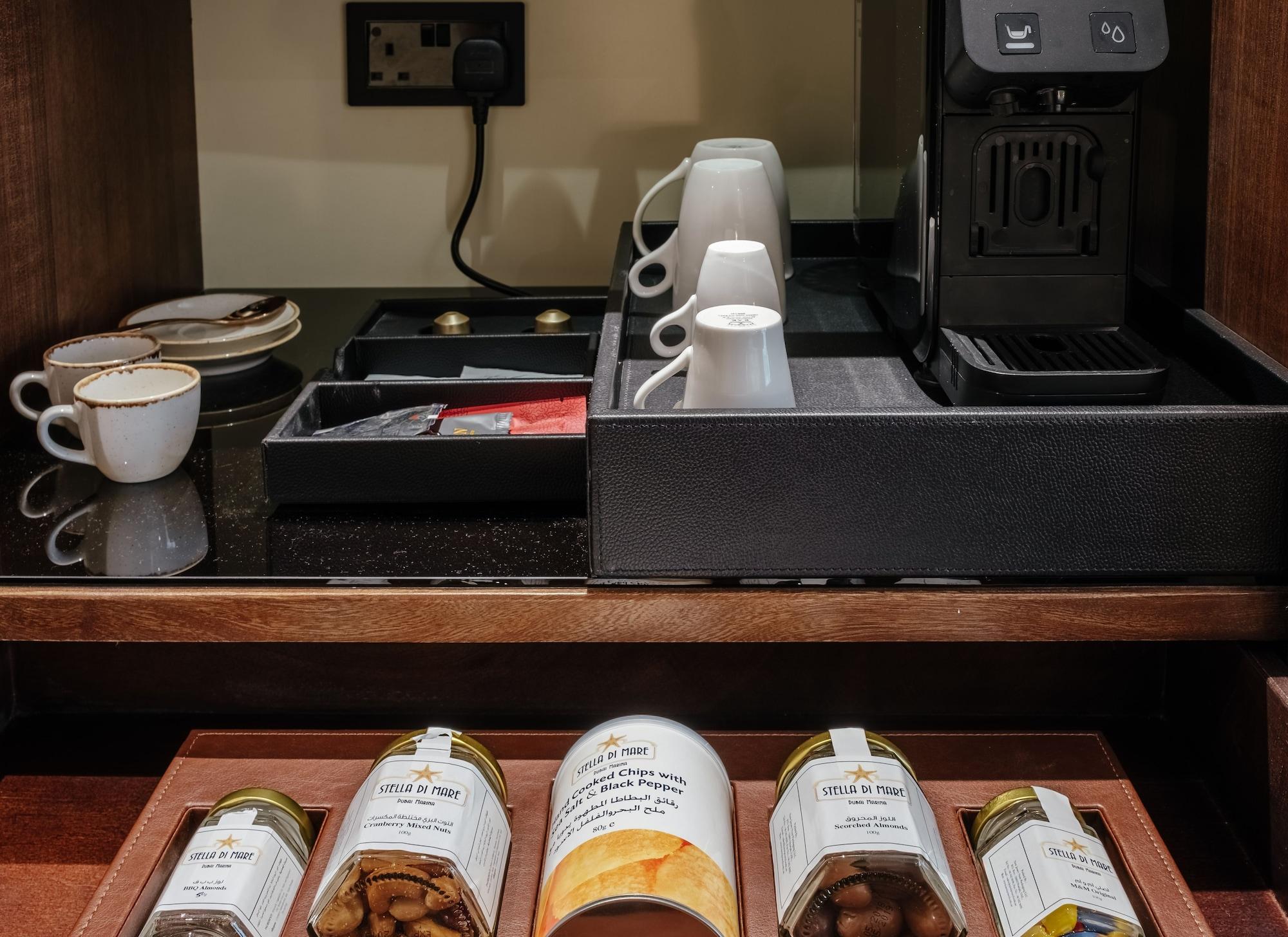 Stella Di Mare Dubai Marina Hotel Esterno foto The photo shows a hospitality setup, likely in a hotel room. It features a countertop with a coffee machine positioned next to several cups and a tray containing various packets. Below, there are jars and a can, presumably containing snacks or condim