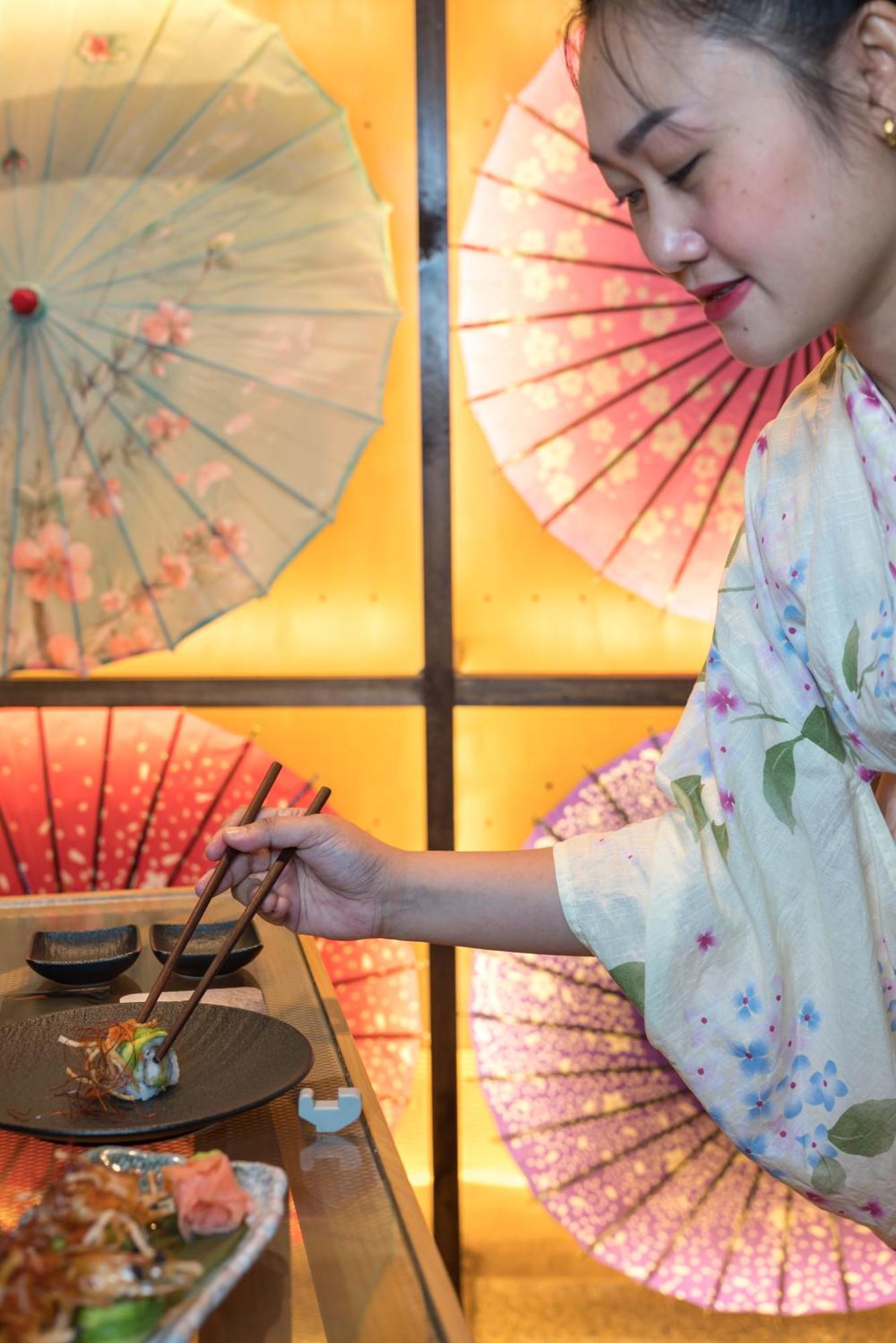 Stella Di Mare Dubai Marina Hotel Esterno foto The photo shows a person wearing a light-colored kimono with floral patterns, holding chopsticks. They are in the process of serving or eating food, which is likely placed on a wooden tray in front of them. Behind them, there are decorative paper umb