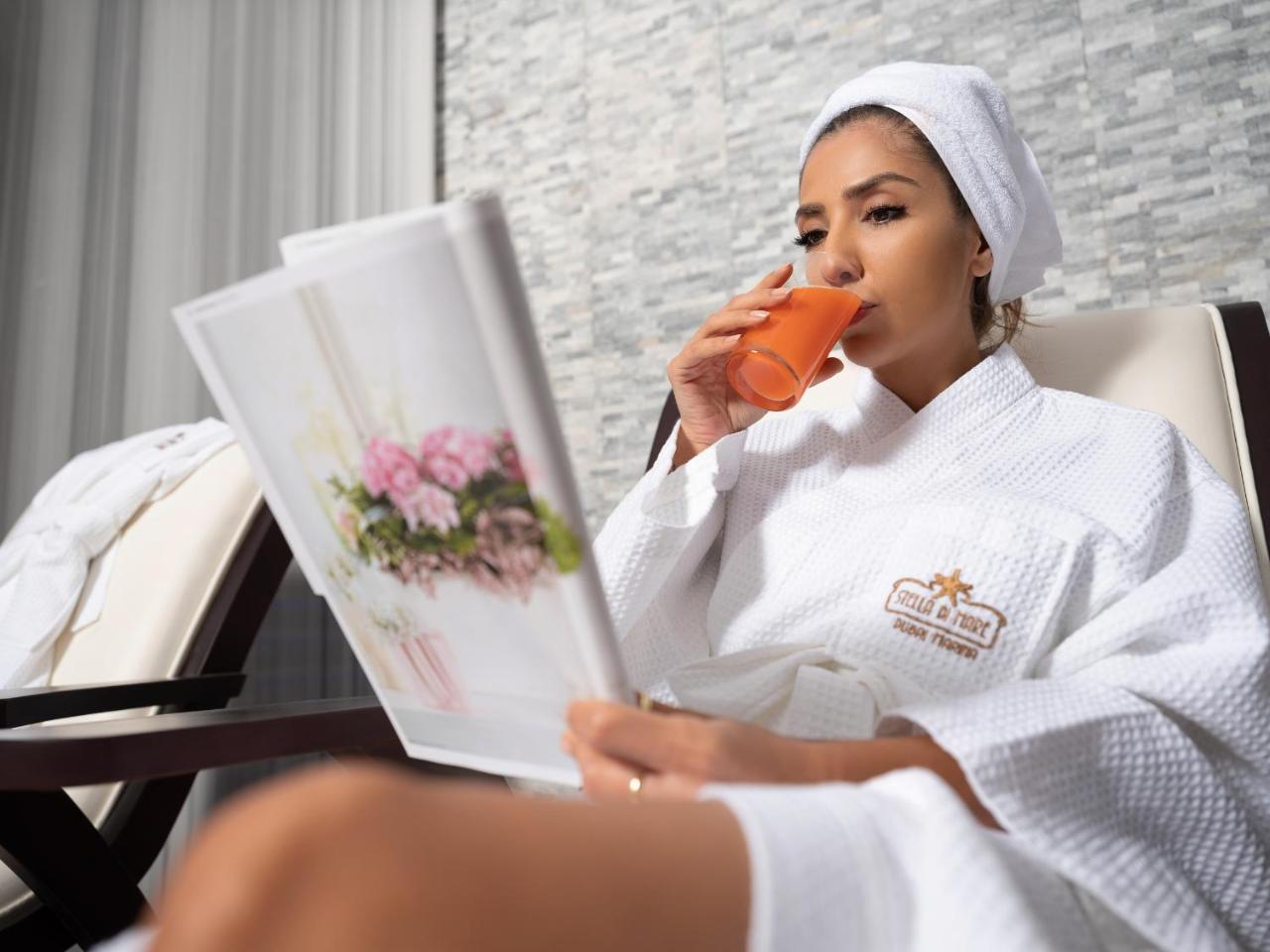Stella Di Mare Dubai Marina Hotel Esterno foto The photo shows a woman sitting in a comfortable setting, likely a spa or relaxation area. She is wearing a white bathrobe and a towel on her head. In one hand, she holds a glass of orange juice, and in the other, she is reading a magazine or brochur