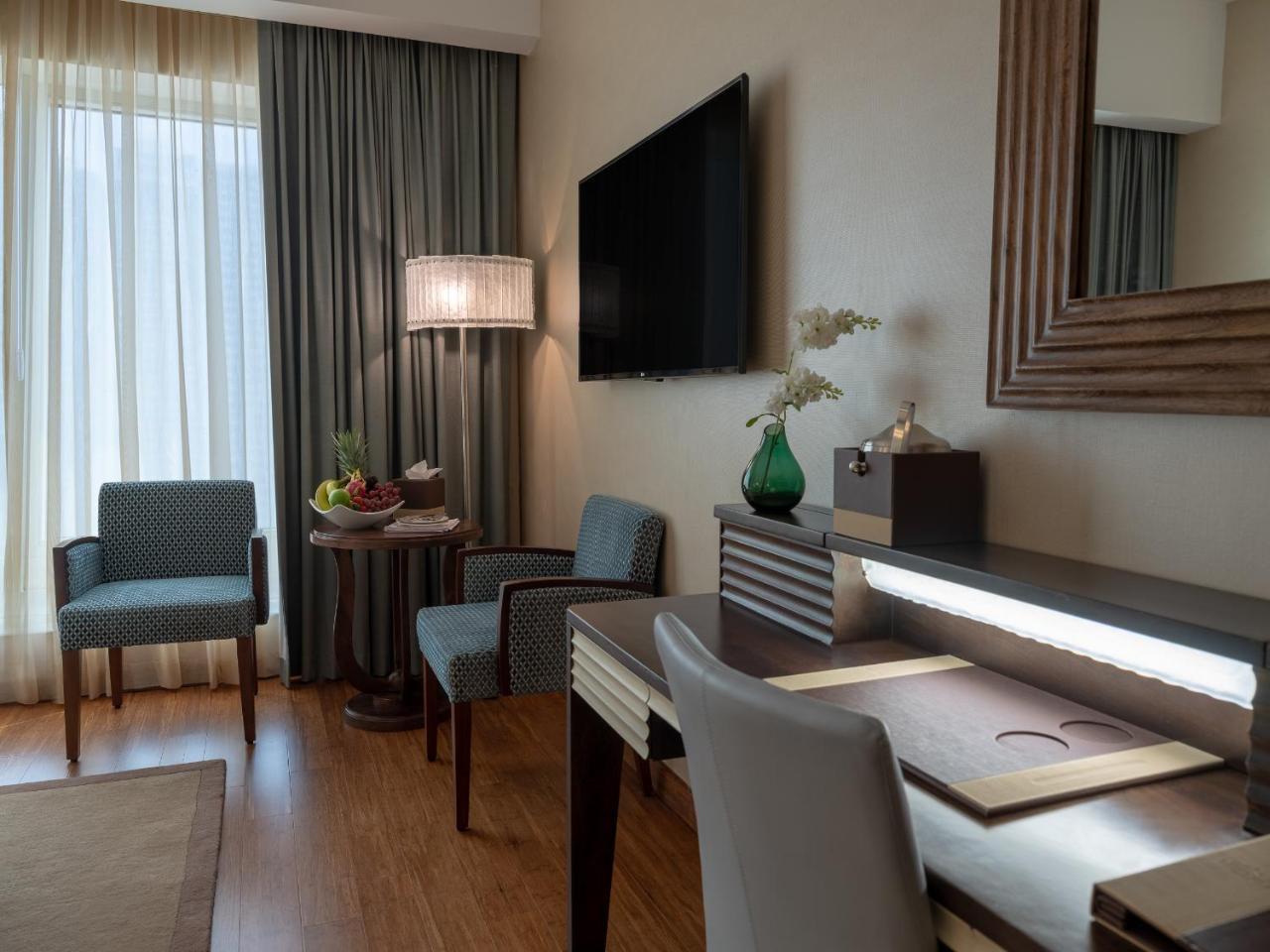 Stella Di Mare Dubai Marina Hotel Esterno foto The photo shows a modern hotel room interior. There's a cozy seating area with two blue chairs and a small round table, which has a fruit arrangement on it. A large window with light curtains allows natural light to fill the room. Opposite the seatin
